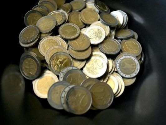 Eine Schüssel mit goldfarbenen Euro-Münzen verschiedener Nennwerte, auf schwarzem Hintergrund präsentiert.