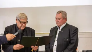 Zwei Männer stehen vor einem Mikrofon und einem Podium. Ein Mann trägt eine Brille und hält ein Buch. Der andere Mann trägt ein weißes Hemd und eine schwarze Jacke.