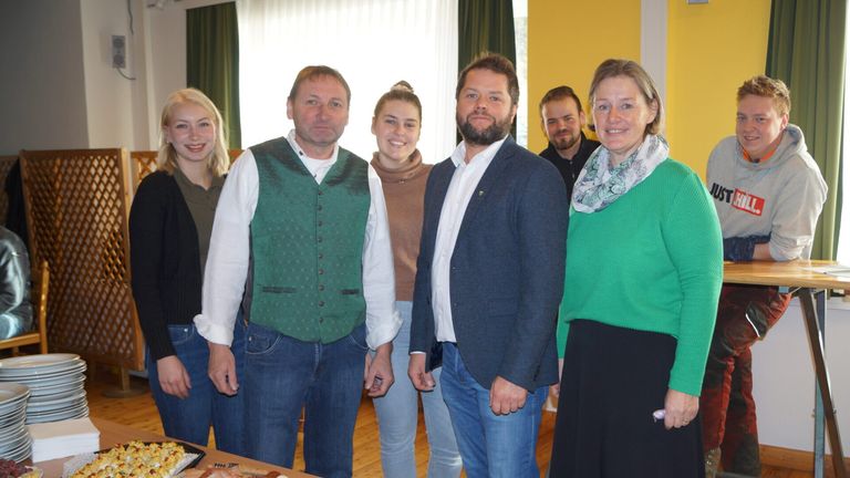 Eine Gruppe von Menschen steht zusammen, wahrscheinlich in einer Besprechung. Ein Mann mit Bart trägt einen blauen Mantel. Die Frau neben ihm trägt einen grünen Pullover und eine Schürze. Die anderen Leute stehen hinter ihnen. Sie alle lächeln. Es gibt einen Tisch mit etwas Essen darauf.