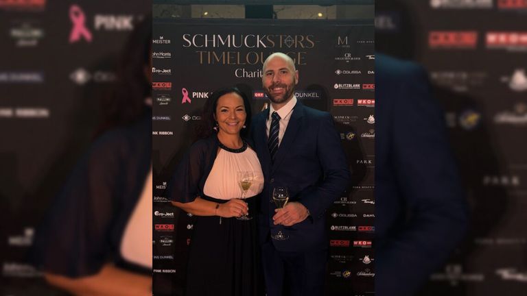 Ein lächelnder Mann und eine Frau, elegant gekleidet, stehen bei einer Wohltätigkeitsveranstaltung zusammen und halten Weingläser. Der Hintergrund zeigt ein rosa Band und verschiedene Logos.