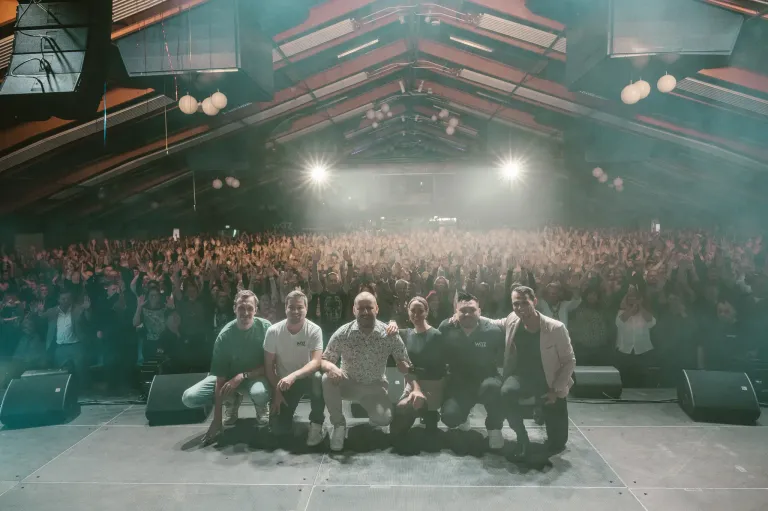 Eine Gruppe von Künstlern posiert auf der Bühne vor einem großen Publikum, während helle Bühnenlichter die Szene beleuchten.