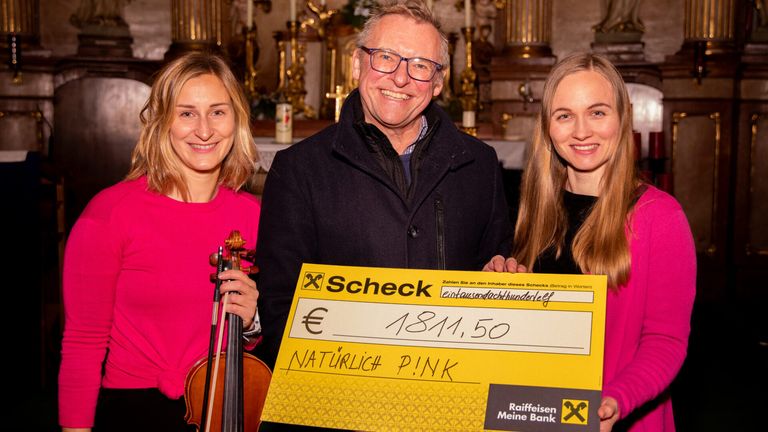 Drei Personen stehen lächelnd eng beieinander in einer Kirche. Der Mann hält einen großen Scheck über 18.111,50 Euro, während die Frauen eine Geige und einen Geigenbogen halten.