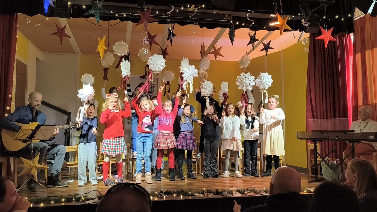 Eine Gruppe von Kindern auf der Bühne, die weiße Papierschneeflocken und rote Bänder halten, mit festlichen Dekorationen darüber.