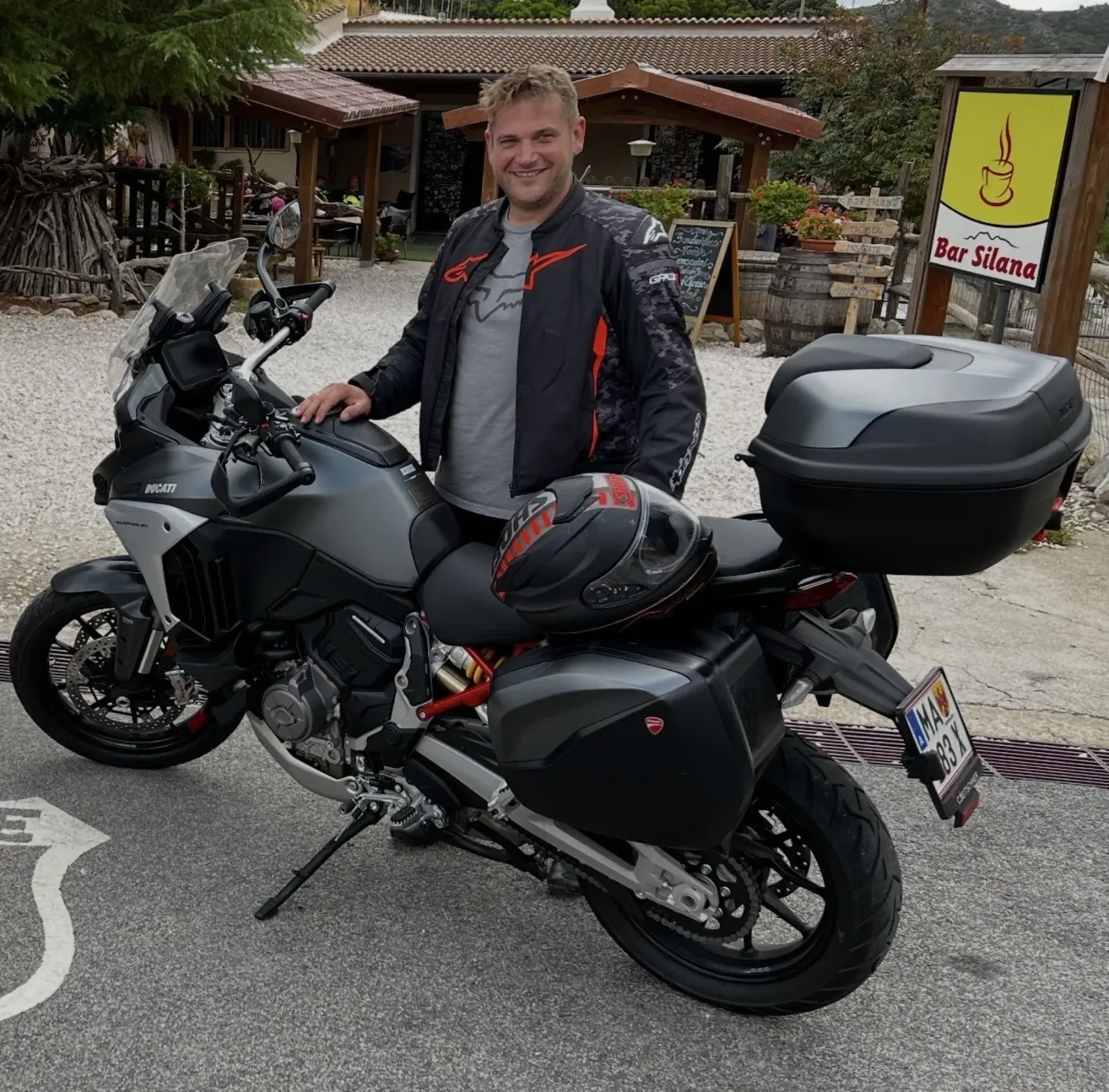 Ein Mann in einer Jacke steht neben einem Ducati-Motorrad. Das Motorrad hat einen schwarzen Helm auf dem Sitz. Im Hintergrund befinden sich ein Gebäude und ein Schild für Bar Silana.