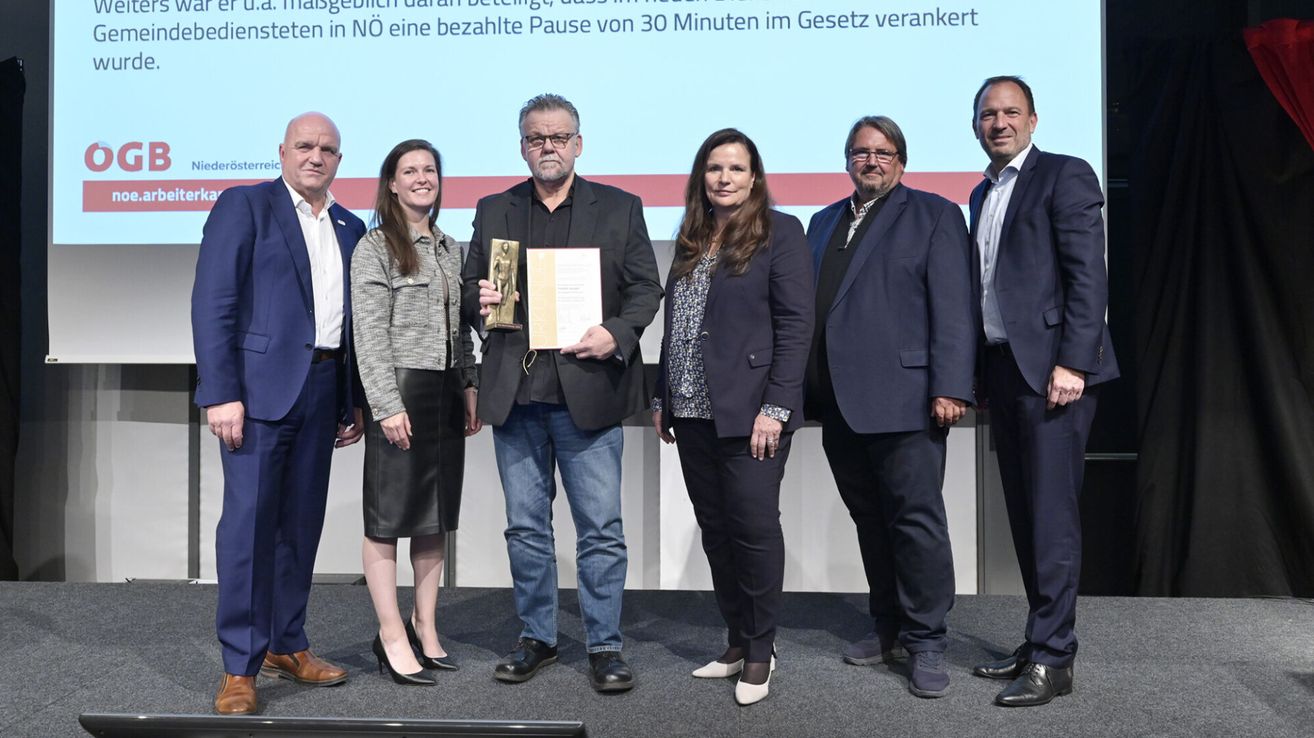 Eine Gruppe von sechs Personen steht auf einer Bühne und hält Auszeichnungen. Ein Mann in der Mitte hält eine goldene Trophäe und ein Zertifikat. Hinter ihnen befindet sich ein Bildschirm mit Text auf Deutsch.