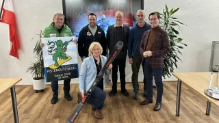 Five men and a woman pose for a photo with skis, a sign, and a large screen in the background.