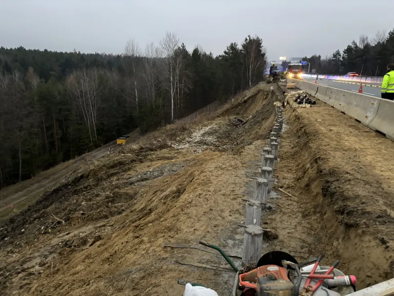 Straße im Bau mit Erde und nahegelegenen Maschinen. Bäume und eine Autobahn sind sichtbar.