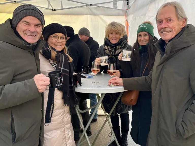 Fünf Menschen lächeln und halten Tassen und Weingläser unter einem Zelt. Sie scheinen sich auf einer gesellschaftlichen Zusammenkunft oder Veranstaltung zu befinden.