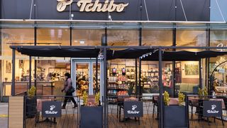 Die Vorderseite eines Tchibo-Ladens mit Glasfassade, einer vorbeigehenden Person und mehreren Außentischen mit Vordächern und Pflanzen in schwarzen Pflanzgefäßen.