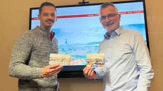 Zwei Männer stehen vor einem Monitor, lächeln und halten Karten mit der Nummer 10 in den Händen. Der Monitor hinter ihnen zeigt eine Stadtlandschaft.