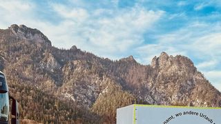 Eine Bergkette mit einem auf der linken Seite geparkten Lkw unter einem teilweise bewölkten Himmel. Die Berge sind felsiger und mit Bäumen bedeckt. Eine weiße Lagerbox mit deutschem Text steht rechts.