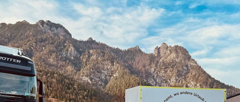 Eine Bergkette mit einem auf der linken Seite geparkten Lkw unter einem teilweise bewölkten Himmel. Die Berge sind felsiger und mit Bäumen bedeckt. Eine weiße Lagerbox mit deutschem Text steht rechts.