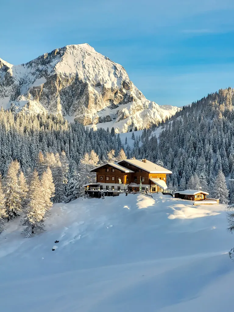 Ein gemütliches Chalet, bedeckt von Schnee, liegt inmitten einer verschneiten Berglandschaft. Hohe Kiefern umgeben das Chalet, und ein größerer schneebedeckter Berg erhebt sich im Hintergrund.