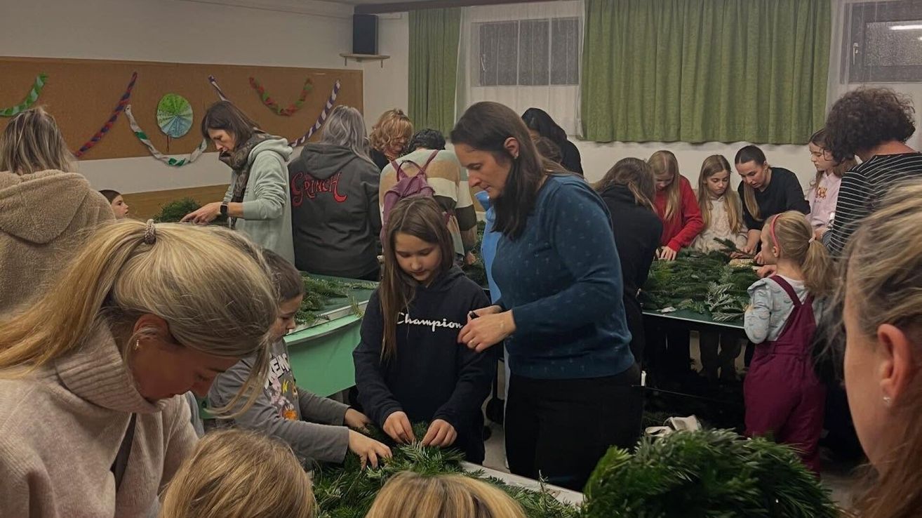 Eine Gruppe von Kindern und Erwachsenen arbeitet gemeinsam in einem Raum, um Kränze zu basteln. Der Raum hat grüne Wände, Vorhänge und einen Lautsprecher. Die Kinder tragen Jacken und einige haben Rucksäcke dabei. Die Kränze bestehen aus Tannenzweigen und haben einige Blumen darauf.