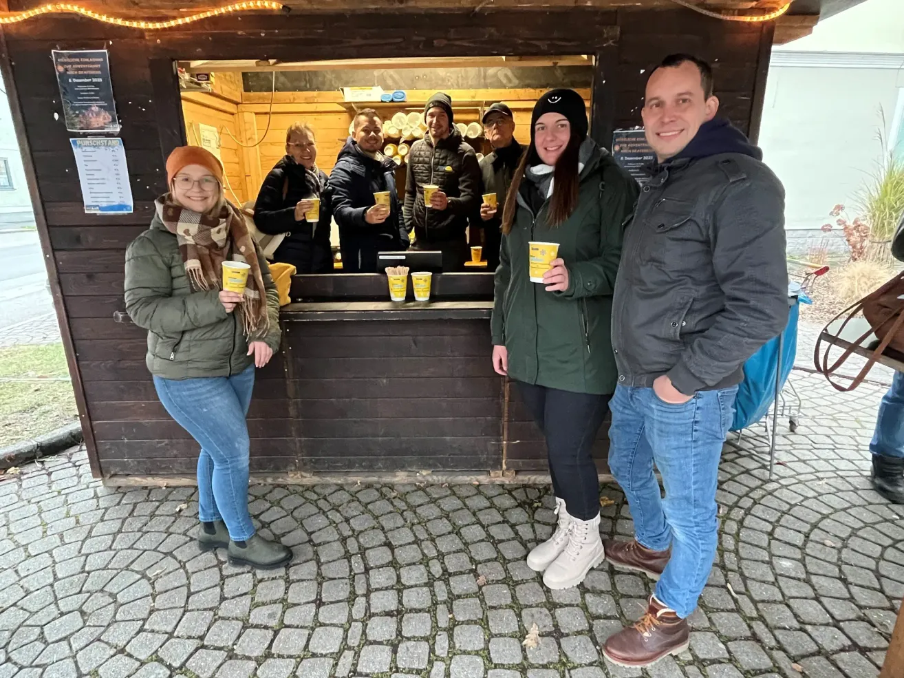 Eine Gruppe von Menschen steht vor einem hölzernen Kiosk und lächelt, während sie Tassen in den Händen halten. Hinter ihnen stehen zwei Frauen und vier Männer im Inneren und halten ebenfalls Tassen. Die Umgebung scheint ein Markt oder ein Festival zu sein.