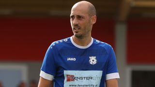 Ein Fußballer in einem blauen Trikot mit weißen Säumen steht auf einem Feld. Das Trikot hat weiße Punkte und das Wort JAKO darauf.