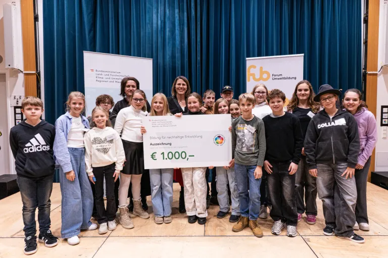 Eine Gruppe von Kindern und Erwachsenen steht hinter einem großen Scheck auf einer Bühne. Sie halten ein Banner und lächeln für die Kamera. Dahinter befinden sich zwei Banner mit Logos und Text.