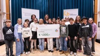 A group of children and adults stand behind a large check on a stage. They are holding a banner and smiling for the camera. Behind them are two banners with logos and text.