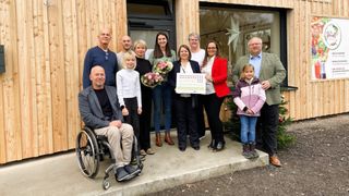 Eine Gruppe von Menschen, darunter ein Mann im Rollstuhl, posiert für ein Foto vor einem Holzgebäude. Sie halten Blumen und ein Schild mit der Aufschrift 'Elsbeere Wienerwald.'