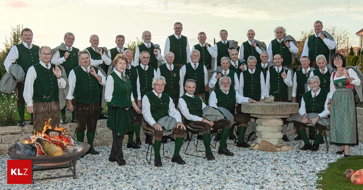 Eine Gruppe älterer Männer und Frauen in traditioneller Kleidung posiert für ein Foto im Garten. Einige sitzen auf Stühlen, andere stehen. Sie alle lächeln und schauen in die Kamera.