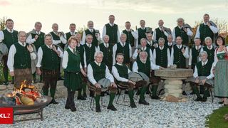 Eine Gruppe älterer Männer und Frauen in traditioneller Kleidung posiert für ein Foto im Garten. Einige sitzen auf Stühlen, andere stehen. Sie alle lächeln und schauen in die Kamera.