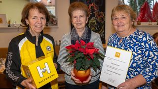 Three smiling women stand together, one holding a certificate, another holding a red poinsettia plant, and the third holding a yellow box with the letter H. Behind them is a wall with a framed picture and a blackboard.