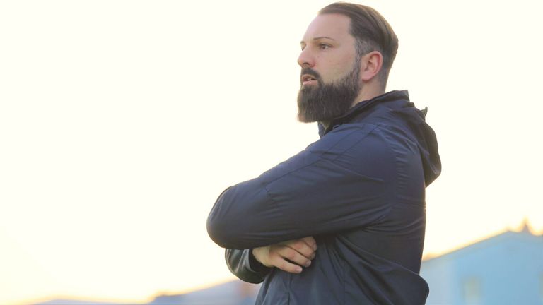 Ein Mann mit Bart steht draußen und trägt eine marineblaue Jacke mit Kapuze. Er hat die Arme vor der Brust verschränkt.