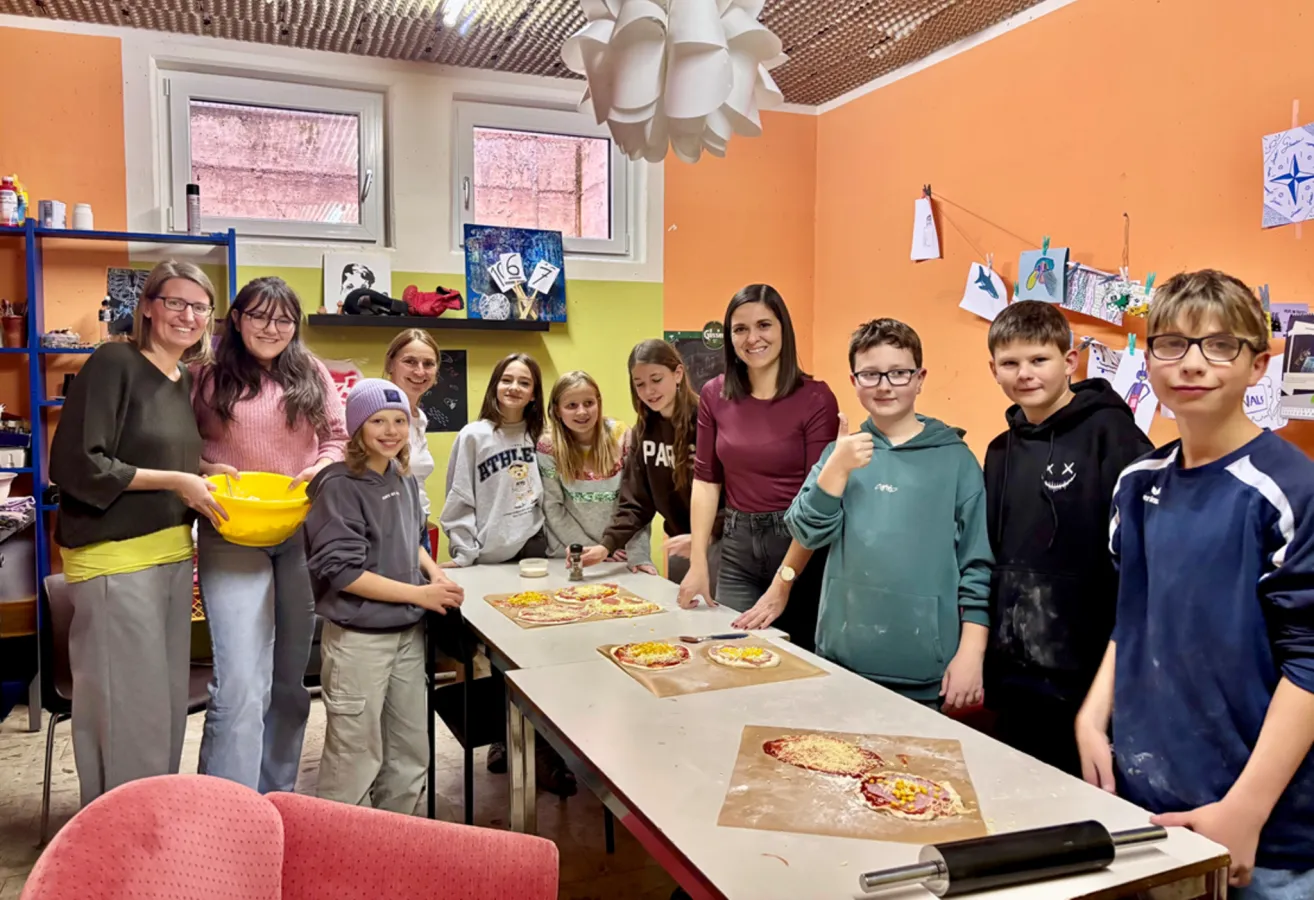 Eine Gruppe von Kindern und Erwachsenen versammelt sich lächelnd um einen Tisch in einem Raum. Der Tisch ist mit Pizzateig und Belägen bestückt. Zwei Frauen stehen lächelnd hinter dem Tisch. Eine Frau hält eine gelbe Schüssel und eine Tafel ist an der Wand.