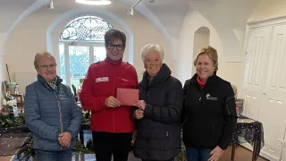 Vier Frauen stehen in einem Raum mit einem Glasfenster, die Frau in Rot hält einen roten Umschlag, die Frau neben ihr hält einen roten Umschlag und die anderen beiden Frauen lächeln.