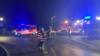 Emergency responders in uniforms and helmets are on a dark street at night. A fire truck with flashing lights is parked nearby. Orange traffic cones mark the area.