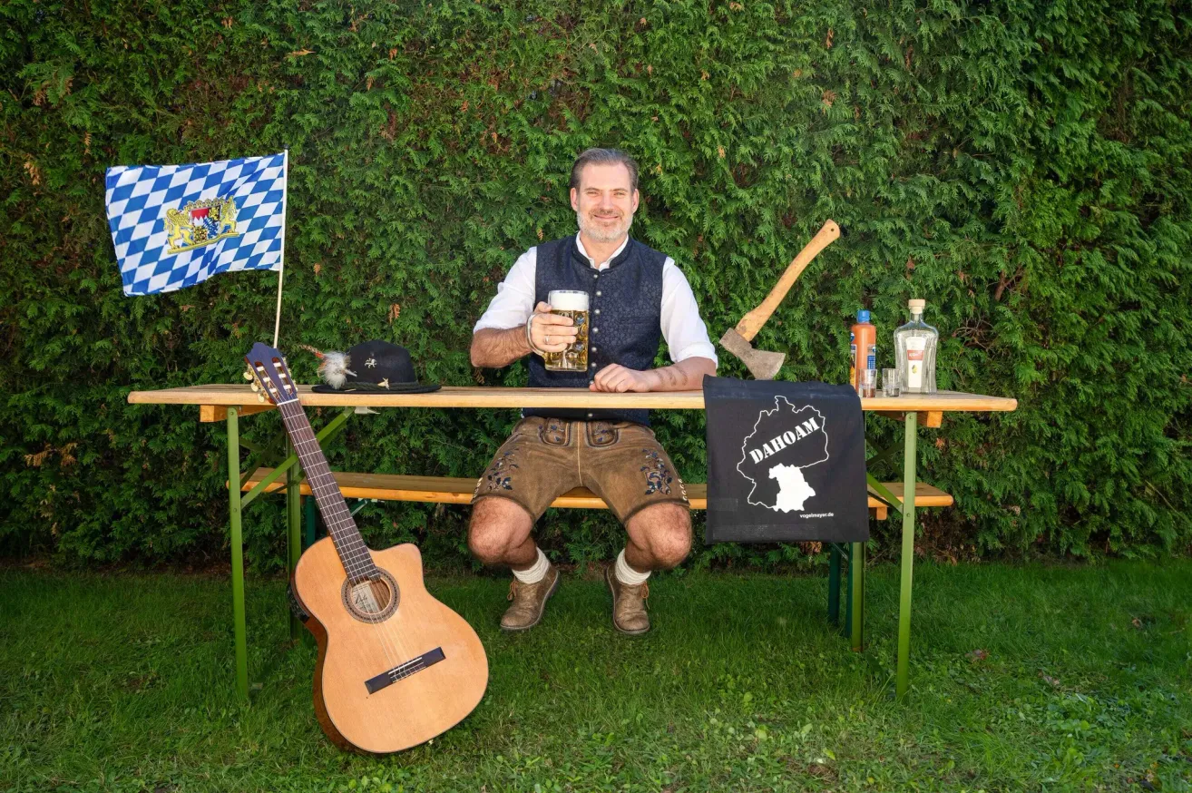 Ein Mann sitzt an einer Bank und hält ein Glas Bier in der Hand, neben ihm steht eine Gitarre. Hinter ihm steht ein Tisch mit einer Flagge, einem Hut, einer Axt, einer Flasche und einem Glas.