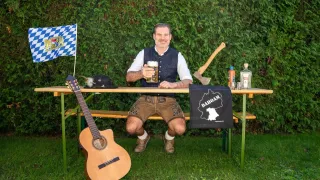 Ein Mann sitzt an einer Bank und hält ein Glas Bier in der Hand, neben ihm steht eine Gitarre. Hinter ihm steht ein Tisch mit einer Flagge, einem Hut, einer Axt, einer Flasche und einem Glas.