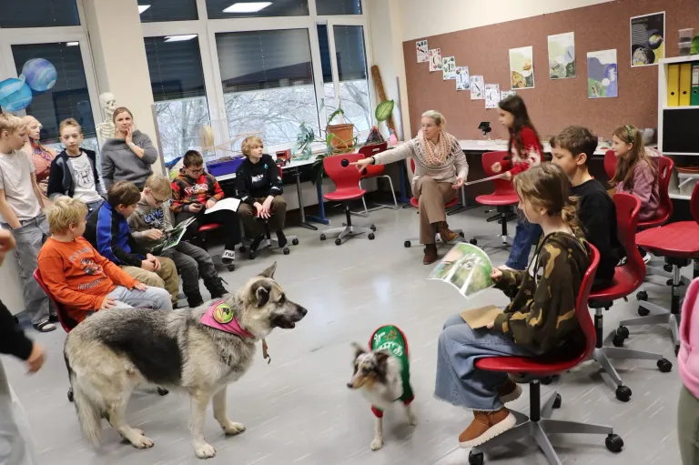 Eine Gruppe von Kindern sitzt auf Stühlen in einem Raum mit zwei Hunden, von denen einer ein grünes Shirt trägt, während eine Frau gestikuliert. Der Raum hat große Fenster und eine Wand mit verschiedenen Postern.