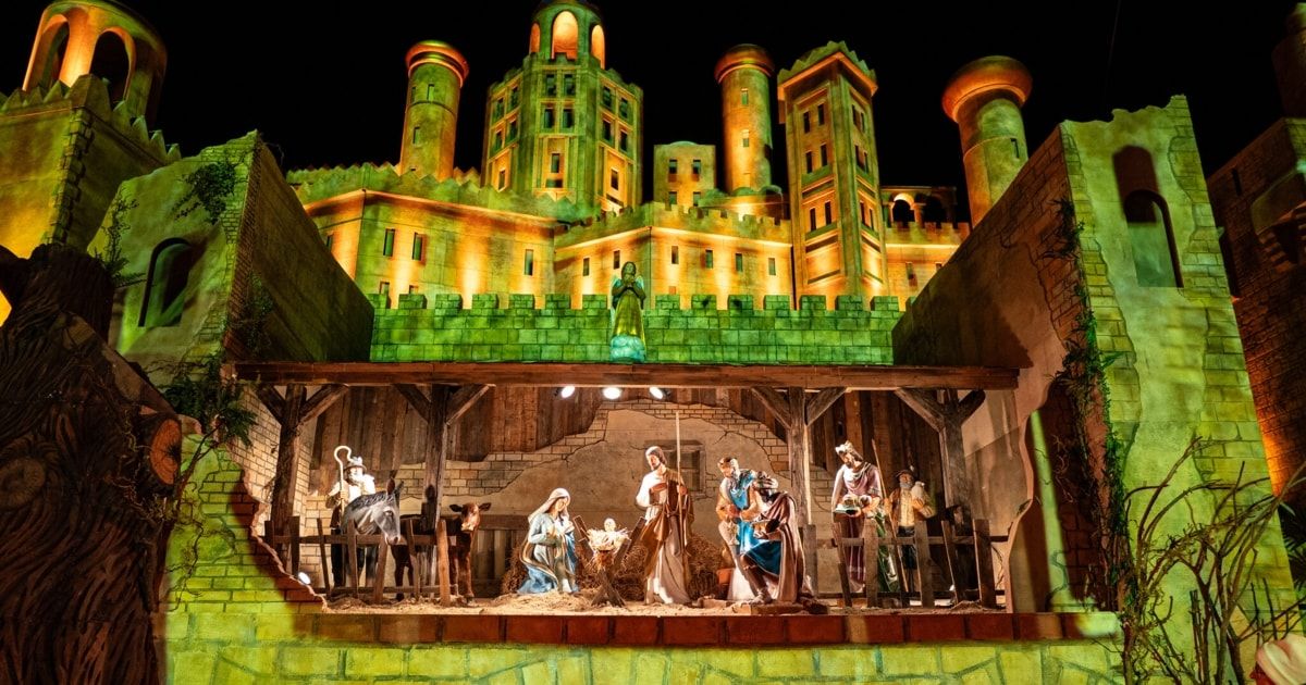 Eine nächtliche Krippe vor einem beleuchteten Schloss mit Türmen. Die Szene zeigt Figuren von Jesus, Maria, Josef und Hirten mit Tieren.