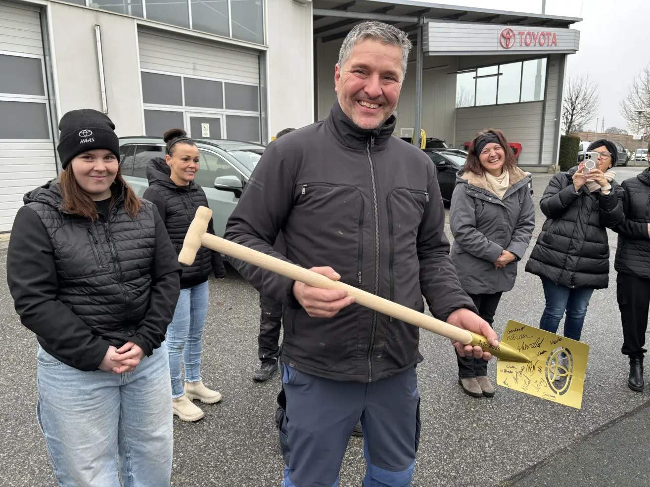 Ein Mann mit einer goldenen Schaufel steht vor einem Toyota-Händler und lächelt, während vier Frauen in Winterkleidung hinter ihm stehen.