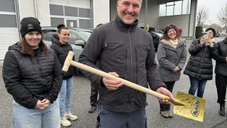 Ein Mann mit einer goldenen Schaufel steht vor einem Toyota-Händler und lächelt, während vier Frauen in Winterkleidung hinter ihm stehen.