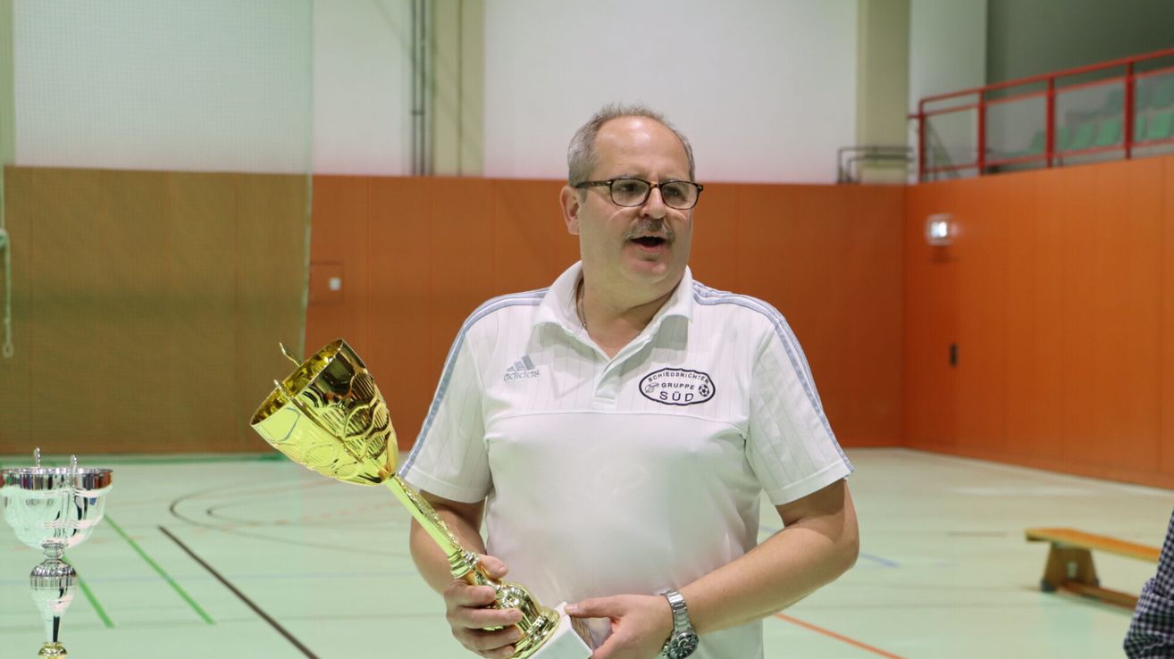 Ein Mann in einem weißen Adidas-Shirt hält eine goldene Trophäe in einer Sporthalle mit orangefarbener Wand.
