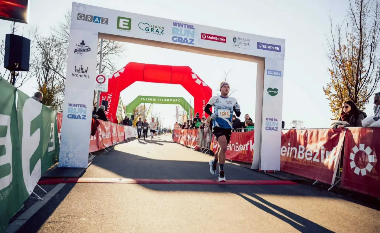 Ein Marathonläufer überquert die Ziellinie beim Grazer Winterlauf, trägt ein blaues Shirt und schwarze Shorts. Hinter ihm jubeln Zuschauer. Im Hintergrund sind Banner und ein roter Bogen zu sehen.
