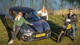 Zwei Frauen stehen neben einem Auto und einem Motorrad auf einer Wiese. Das Auto hat ein gelbes Nummernschild. Eine Frau trägt eine Sonnenbrille und eine Jacke.