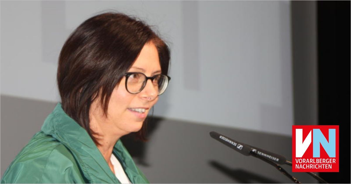 Eine Frau mit Brille und grüner Jacke lächelt und spricht in ein Sennheiser-Mikrofon. Der Hintergrund ist eine schlichte, helle Wand.