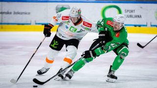 Zwei Eishockeyspieler in verschiedenen Trikots spielen auf der Eisbahn. Einer trägt ein weißes Trikot, der andere ein grünes. Beide halten Eishockeyschläger.