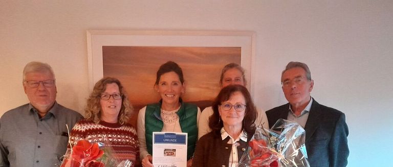 Eine Gruppe von Menschen steht lächelnd zusammen, hält Blumen und ein Zertifikat. Sie sind in einem Raum mit einem gerahmten Gemälde an der Wand.