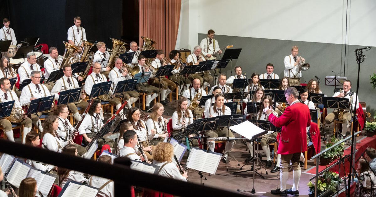 Ein Orchester auf der Bühne mit einer Mischung aus Blechblas- Holzblas- und Perkussionsinstrumenten. Ein Dirigent leitet von vorne, und das Publikum sitzt davor.