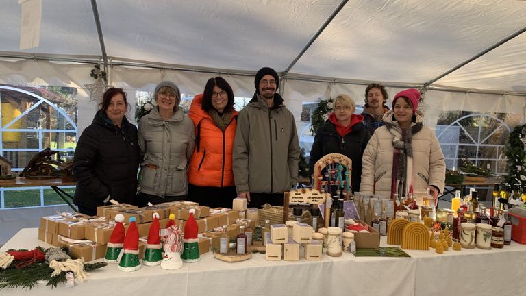 Mehrere Menschen stehen unter einem Zelt und lächeln. Vor ihnen steht ein Tisch voller verschiedener Produkte.