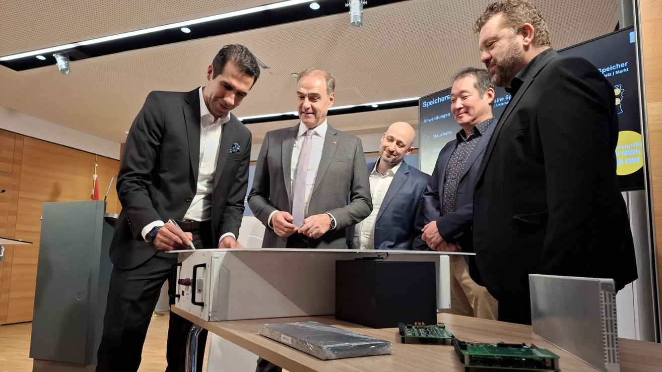 Fünf Männer in Anzügen stehen um einen Tisch mit elektronischer Ausrüstung. Ein Mann schreibt auf einen Clipboard, während ein anderer auf ein Gerät zeigt.