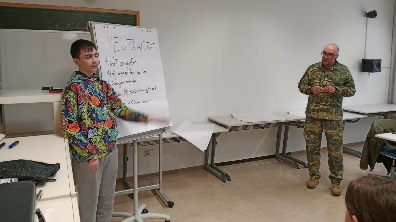 Ein junger Mann in einem farbenfrohen Shirt steht vor einer Tafel mit der Aufschrift 'Neutralität'. Daneben steht eine Person in Militäruniform neben einem Schreibtisch. Papiere liegen auf dem Schreibtisch.