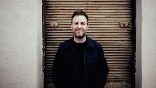 Ein Mann mit Bart und Schnurrbart steht lächelnd vor einer rostigen Metallklappe und posiert für die Kamera. Er trägt eine dunkle Jacke und ein schwarzes Hemd.