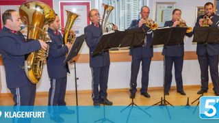 Eine Gruppe von Musikern in Militäruniformen spielt Blechblasinstrumente in einem Raum mit Holzböden und weißen Wänden.