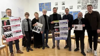 Eine Gruppe von Personen steht drinnen und hält jeweils ein Werbeposter. Sie lächeln und scheinen sich bei einer Veranstaltung zu befinden. Die Poster haben Text und Bilder. An der Wand hinter ihnen sind mehrere Poster angebracht.