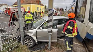 Rettungskräfte in gelben Jacken inspizieren einen silbernen Wagen, der mit einem Pfosten kollidiert ist. Die Front des Autos ist schwer beschädigt. Ein Feuerwehrmann steht mit orangem Helm daneben.